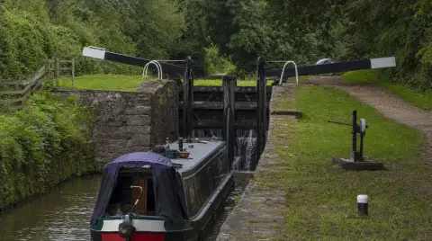 A narrowboat waiting below a canal lock, the black and white balance beams and paddle gear on the lockside.