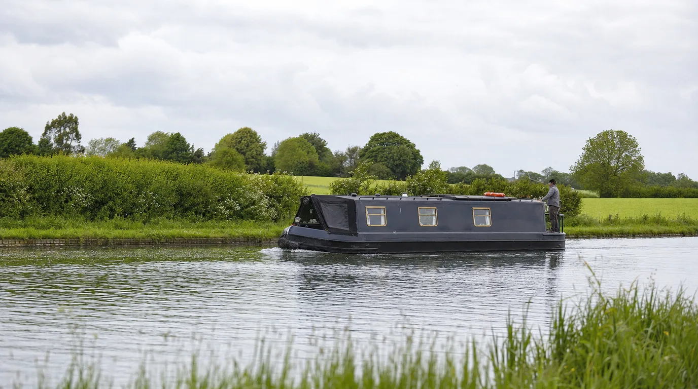 A narrowboat cruising a wide, quiet reach of canal, the steerer standing at the stern.